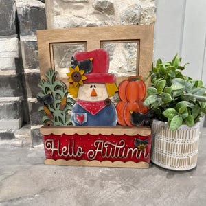 May include: A wooden fall-themed decoration featuring a scarecrow with a red hat, blue shirt, and a red bandana. The decoration includes a pumpkin, a sunflower, and the text "Hello Autumn" on a red base. A small potted plant is to the right.