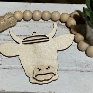 May include: A wooden cutout of a cow's head with a striped pattern on the forehead. The cutout is painted white and has a natural wood grain texture. The cow's head is hanging from a string of wooden beads.