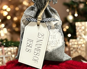 May include: A gray burlap gift bag with a drawstring closure, holding small items. A white tag is attached with the text "25 Names of Jesus" and "The Christ". The bag sits on a red, textured surface.