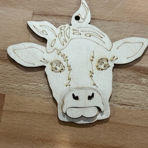 May include: A wooden cutout of a cow's head with a simple design. The cow has large eyes, a wide nose, and a tongue sticking out.