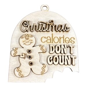 May include: A light-coloured wooden ornament shaped like a dome with a hanging hole at the top. It features a layered gingerbread man figure with three small buttons and the text "Christmas calories DON'T COUNT" engraved in a mix of script and block fonts.