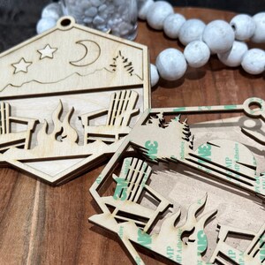 May include: Two wooden cutouts of a campfire scene with two chairs and a moon and stars. The cutouts are unfinished and have a brown wood color. The cutouts are on a brown wooden surface.