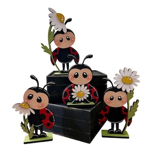 May include: Four wooden ladybug figurines with black bodies, red spotted wings, and smiling faces. Each ladybug holds a white daisy with a yellow center. The figurines are on small green bases and arranged on a black block.