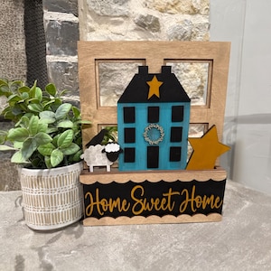 May include: A wooden decorative piece featuring a blue house with a black roof and a gold star. A sheep and a gold star are next to the house. The words "Home Sweet Home" are written in gold script on a black background.