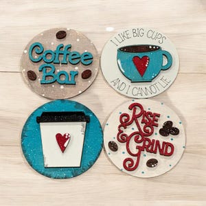 May include: Four round wooden signs with coffee-themed sayings. The signs are painted in shades of brown, white, and blue. The text on the signs reads "Coffee Bar", "I like big cups and I cannot lie", "Rise & Grind", and a coffee cup with a heart.