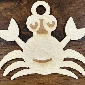 May include: A wooden cutout of a crab with a simple, cartoon-like face. The crab is light brown and has a hole at the top for hanging.