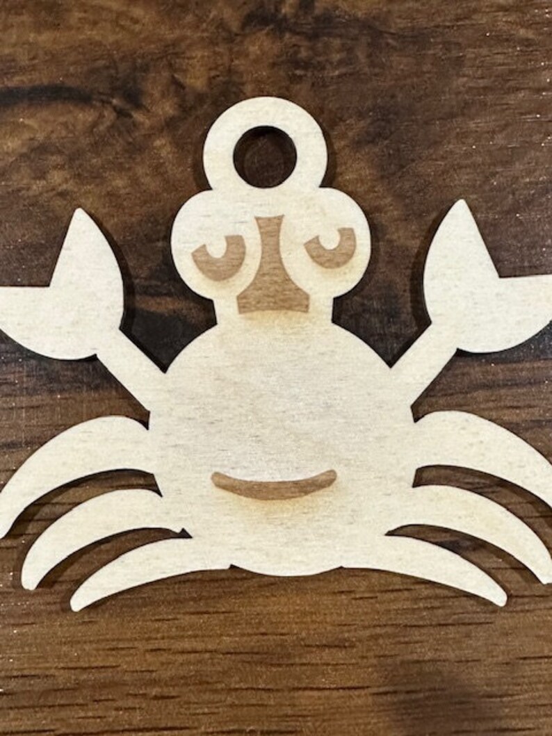 May include: A wooden cutout of a crab with a simple, cartoon-like face. The crab is light brown and has a hole at the top for hanging.