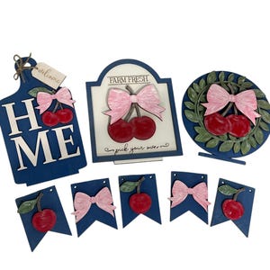 May include: A collection of blue wooden cherry-themed decorations. Includes a cutting board with "HOME" and "ME", a sign that says "FARM FRESH", a wreath, and several pennant banners. Each piece features red cherries and pink bows.