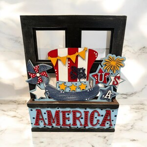 May include: A wooden window frame with a red, white, and blue patriotic hat, stars, and fireworks inside. The window frame has a wooden box below it with the word "AMERICA" in red letters on a blue background.