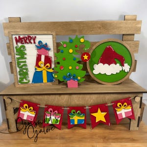 May include: Wooden Christmas decorations on a wooden bench. Includes a framed sign with "Merry Christmas" and stacked gifts, a green Christmas tree, and a round sign with a Santa hat. A banner of gift-shaped flags hangs below.