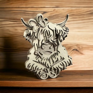 May include: A wooden cutout of a Highland cow with a bow on its head. The cow is sitting with its legs tucked under its body. The cutout is made of light brown wood and has a detailed design.