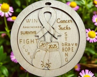 May include: A wooden round ornament with a pink ribbon for hanging. The ornament features a ribbon, boxing gloves, and the words "WIN", "SURVIVE", "FIGHT", "CURE", "Cancer Sucks", "bRAVE", "HOPE", and "20 2025".