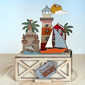 May include: A multi-layered wooden decorative display featuring a lighthouse, a sailboat, a sandcastle, palm trees, and a sun. The base is a rectangular box with a light blue tag attached that reads Ocean ESCAPE in orange and dark gray lettering.