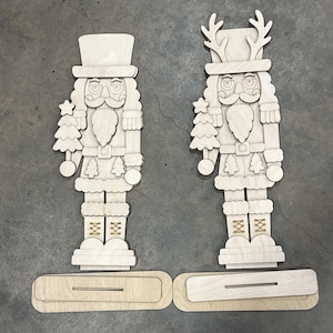 May include: Two unfinished light-colored wooden nutcracker cutouts. One nutcracker wears a top hat and holds a small tree, while the other features antlers on its hat and holds a tree. Each figure includes a separate rectangular wooden base.