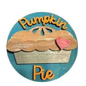 May include: A round wooden sign with a blue background and a cutout of a pumpkin pie with a red heart on top. The words "Pumpkin Pie" are written in orange letters on the sign.