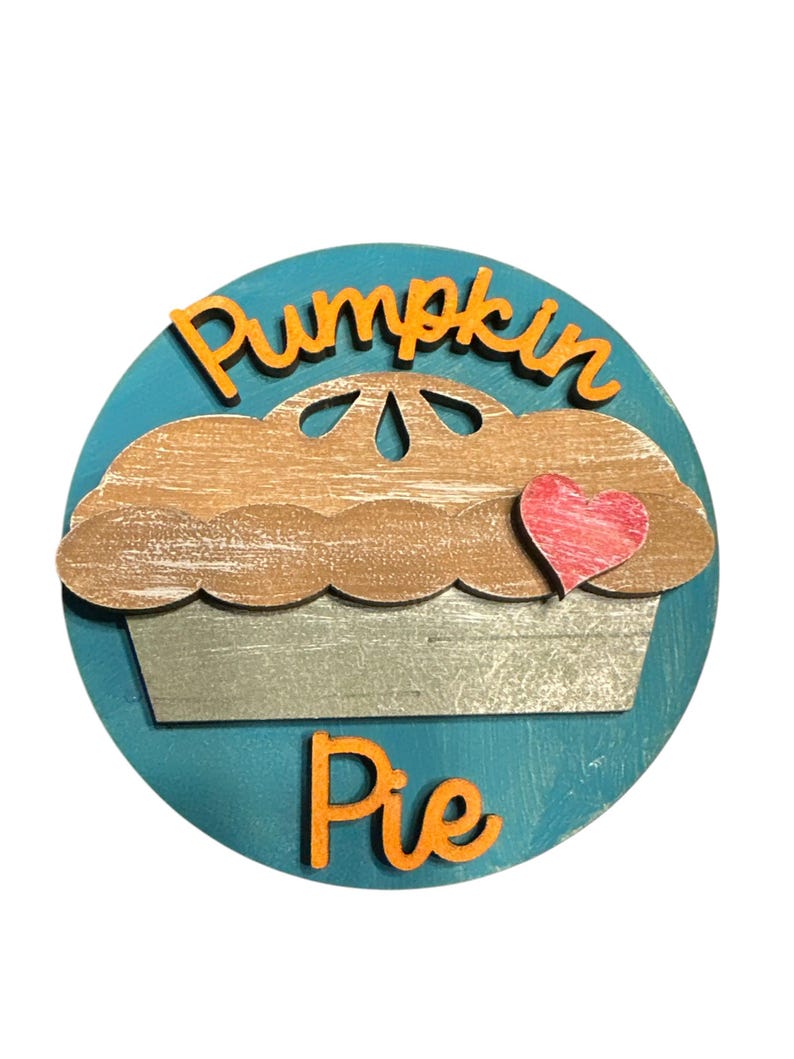 May include: A round wooden sign with a blue background and a cutout of a pumpkin pie with a red heart on top. The words "Pumpkin Pie" are written in orange letters on the sign.