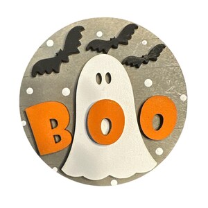 May include: A round wooden sign with a gray background and white polka dots. A white ghost with orange letters spelling "BOO" is in the center. Black bats are flying around the ghost.