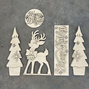 May include: A set of five light-colored wooden laser-cut decorations including two pine trees with star accents, a reindeer with a bow and bell, a circular sign with the text Merry & Bright, and a rectangular sign with the text Seasons Greetings.