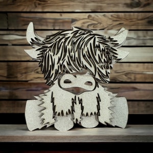 May include: A wooden cutout of a Highland cow with a shaggy brown and black coat. The cow is sitting with its legs tucked under its body and has a friendly expression.