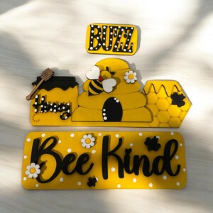 May include: Wooden bee-themed sign with the words "Buzz", "Honey", and "Bee Kind". The sign features a beehive, a bee, a honey pot, and white polka dots.