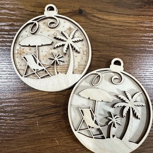 May include: Two wooden ornaments shaped like circles with a beach scene. The ornaments feature palm trees, lounge chairs, and umbrellas. The ornaments are unfinished and ready to be painted or stained.