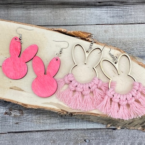 May include: Two pairs of pink bunny-shaped earrings. One pair is solid pink wood, the other pair is natural wood with pink macrame fringe.