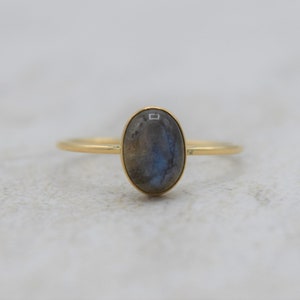 May include: A gold ring featuring an oval-shaped labradorite gemstone. The stone has a mix of gray and blue hues. The ring's band is thin and made of gold. The ring is a piece of jewelry.