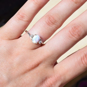 May include: A silver ring with an oval opal center stone and two smaller accent stones. The ring has a delicate, swirling design. The opal has a milky white and iridescent appearance. Jewelry.