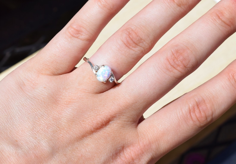 May include: A silver ring with an oval opal center stone and two smaller accent stones. The ring has a delicate, swirling design. The opal has a milky white and iridescent appearance. Jewelry.