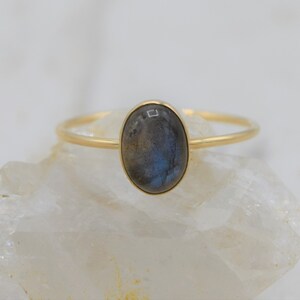 Puede incluir: Anillo de oro con una piedra labradorita ovalada. El anillo tiene una banda fina y un engaste de bisel. La piedra muestra tonos grises, azules y verdes. El anillo se exhibe sobre una superficie blanca y texturizada. La piedra mide aproximadamente 1,5 cm.