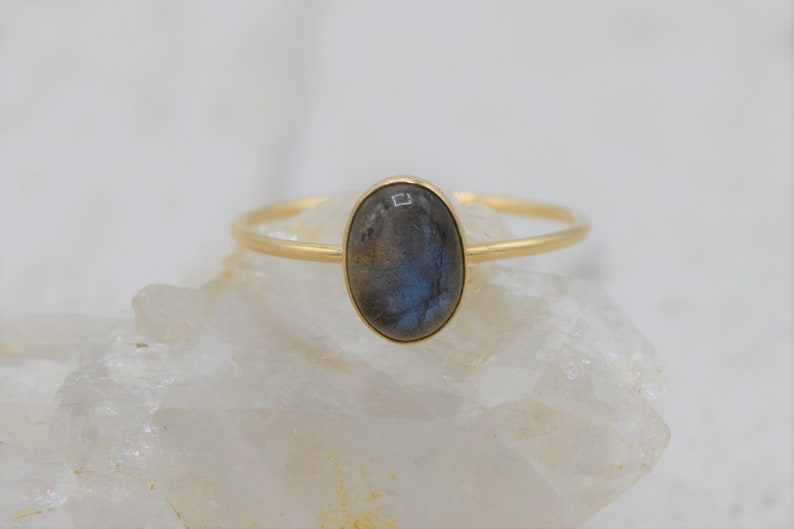 Puede incluir: Anillo de oro con una piedra labradorita ovalada. El anillo tiene una banda fina y un engaste de bisel. La piedra muestra tonos grises, azules y verdes. El anillo se exhibe sobre una superficie blanca y texturizada. La piedra mide aproximadamente 1,5 cm.