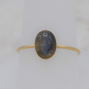 Puede incluir: Un anillo de oro con una piedra preciosa ovalada e iridiscente. La piedra est&aacute; engastada en un bisel de oro sobre una fina banda de oro. El anillo se muestra sobre un fondo blanco, resaltando su dise&ntilde;o elegante y atemporal.