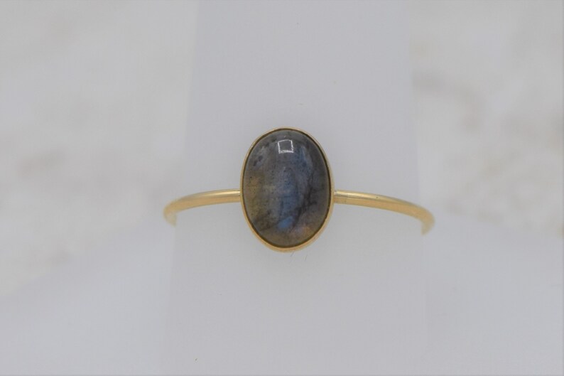 Puede incluir: Un anillo de oro con una piedra preciosa ovalada e iridiscente. La piedra est&aacute; engastada en un bisel de oro sobre una fina banda de oro. El anillo se muestra sobre un fondo blanco, resaltando su dise&ntilde;o elegante y atemporal.