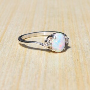 May include: A silver ring with an oval, iridescent opal center stone and two smaller clear stones on either side. The ring is set on a light wooden surface.