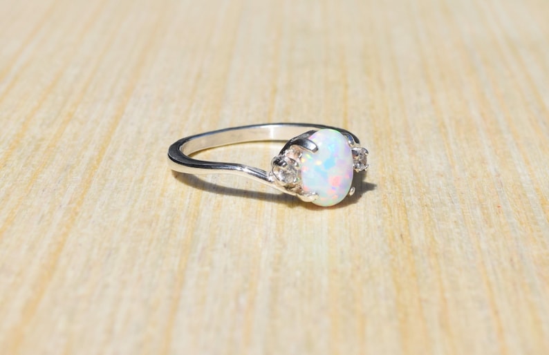 May include: A silver ring with an oval, iridescent opal center stone and two smaller clear stones on either side. The ring is set on a light wooden surface.