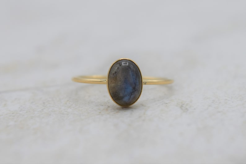 Puede incluir: Un anillo de oro con una piedra preciosa de labradorita ovalada. La piedra tiene tonos grises y azules. El anillo tiene una fina banda de oro. Es una joya.