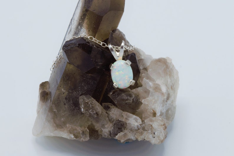 Puede incluir: Un colgante de &oacute;palo blanco en una cadena de plata, descansando sobre un grupo de cristales de cuarzo ahumado.
