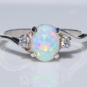 May include: A silver ring featuring an oval opal gemstone with iridescent color flashes. Two smaller, round, clear gemstones flank the opal. The ring has a delicate, swirling band design, reflecting light.