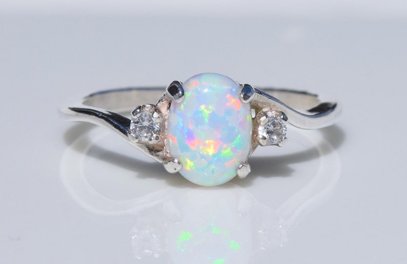 May include: A silver ring featuring an oval opal gemstone with iridescent color flashes. Two smaller, round, clear gemstones flank the opal. The ring has a delicate, swirling band design, reflecting light.