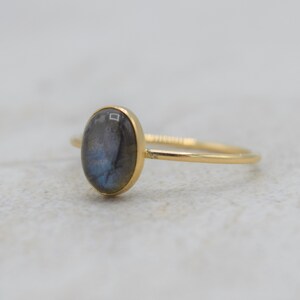 Puede incluir: Anillo de oro con una piedra de labradorita ovalada. La piedra muestra tonos grises y azules. El anillo tiene una banda fina. La joya est&aacute; sobre un fondo neutro. Una joya elegante y discreta.