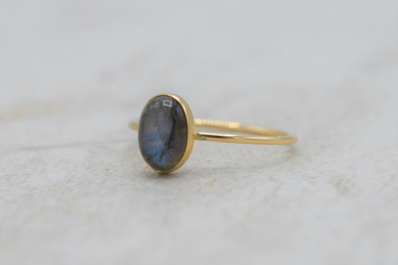 Puede incluir: Anillo de oro con una piedra de labradorita ovalada. La piedra muestra tonos grises y azules. El anillo tiene una banda fina. La joya est&aacute; sobre un fondo neutro. Una joya elegante y discreta.