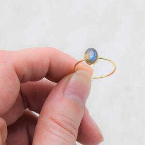 Puede incluir: Un delicado anillo de oro con una piedra preciosa de labradorita de forma ovalada. La fina banda del anillo resalta los tonos iridiscentes azules y amarillos de la piedra. Una joya minimalista.