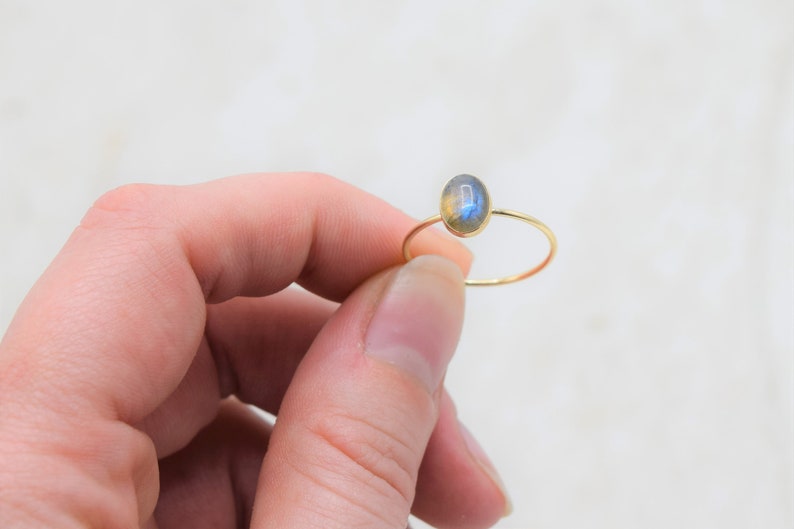 Puede incluir: Un delicado anillo de oro con una piedra preciosa de labradorita de forma ovalada. La fina banda del anillo resalta los tonos iridiscentes azules y amarillos de la piedra. Una joya minimalista.
