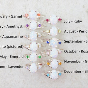 May include: A collection of silver rings, each featuring a large oval white gemstone and smaller accent stones in various colors. Text indicates each ring represents a birthstone for a month, including Garnet, Amethyst, Emerald, and Ruby.
