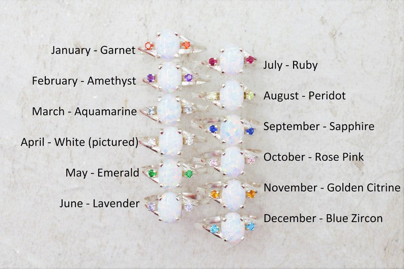May include: A collection of silver rings, each featuring a large oval white gemstone and smaller accent stones in various colors. Text indicates each ring represents a birthstone for a month, including Garnet, Amethyst, Emerald, and Ruby.