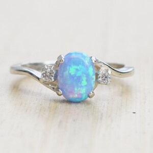 May include: A silver ring with a blue opal gemstone and two smaller clear gemstones. The ring has a wavy band.