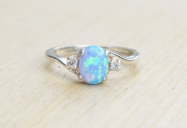 May include: A silver ring with a blue opal gemstone and two smaller clear gemstones. The ring has a twisted band.
