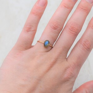 Puede incluir: Un anillo de oro con una peque&ntilde;a piedra preciosa ovalada azul y verde. El anillo est&aacute; en un dedo, con la mano visible. La banda del anillo es delgada y delicada.
