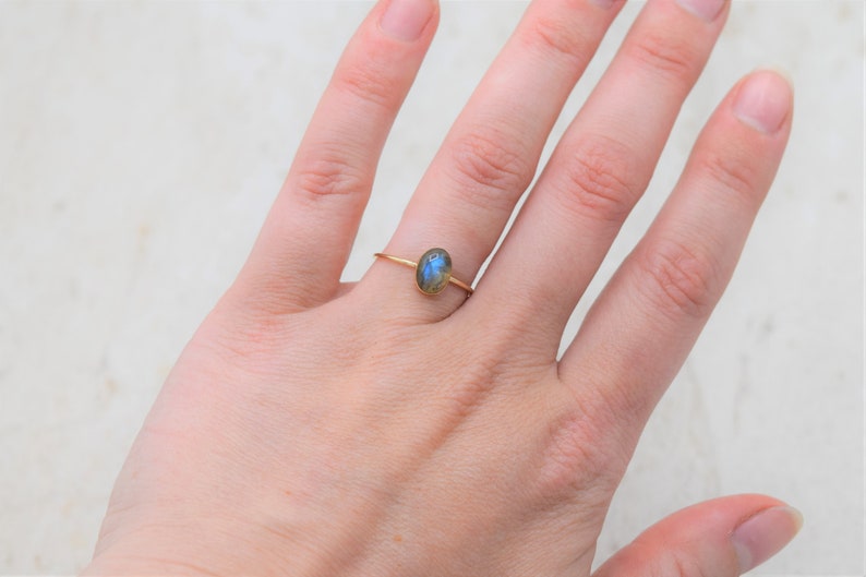 Puede incluir: Un anillo de oro con una peque&ntilde;a piedra preciosa ovalada azul y verde. El anillo est&aacute; en un dedo, con la mano visible. La banda del anillo es delgada y delicada.