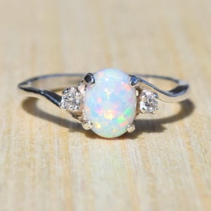 May include: A silver ring featuring an oval, iridescent opal center stone. Two smaller, round, clear gemstones flank the opal. The ring's band has a swirling design. The ring is set against a light wood grain background.